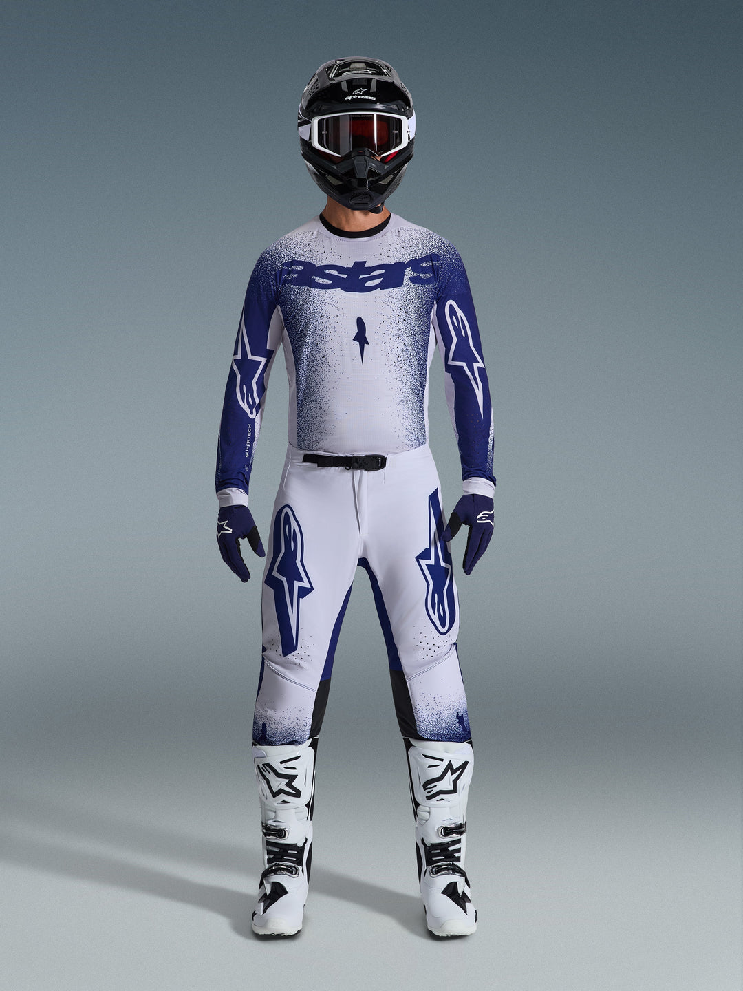 Motocross rider wearing the Alpinestars 2026 Supertech Scenz Gear Set, white and dark blue jersey with matching pants, features large Astars logos and speckled graphic design, paired with a black helmet, white and black protective boots, and coordinated blue gloves, complete professional racing outfit