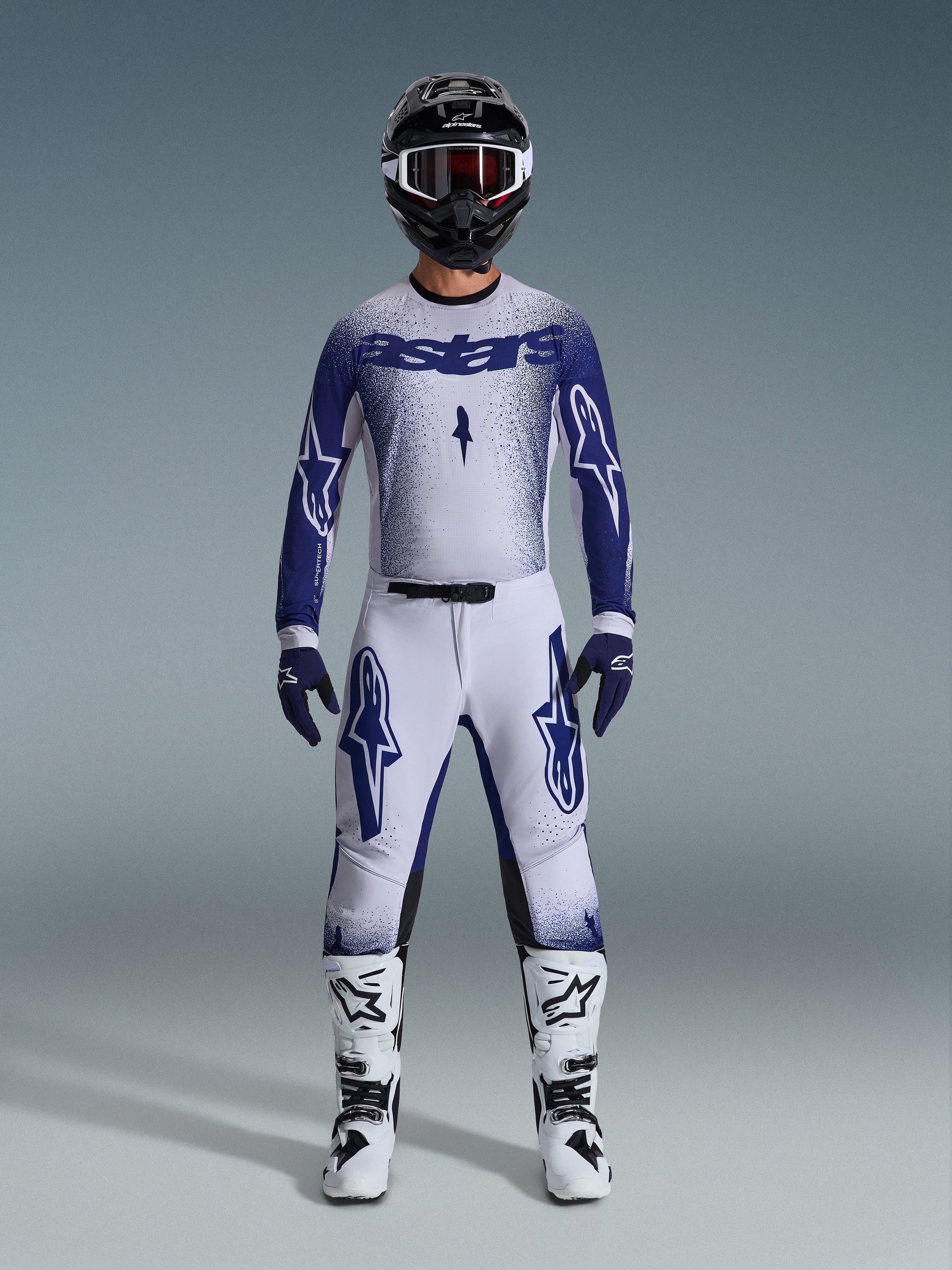 Motocross rider wearing Alpinestars 2026 Supertech Scenz gear set, Light Gray and Dark Blue, including a jersey and pants with matching blue gloves, white and black boots, and a black helmet with white goggles, coordinated outfit for professional off-road racing