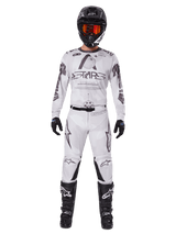 Motocross rider wearing the Alpinestars 2025 Racer Hollow Pants in Light Camo Brown, paired with a matching white and grey long-sleeve jersey, black helmet with orange-red goggles, black gloves, and black boots, coordinated racing gear set for off-road performance.