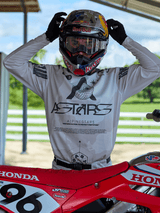Motocross rider wearing Alpinestars 2025 Racer Hollow gear, light camo jersey with bold ASTARS branding, adjusting a Red Bull helmet with clear goggles, standing next to a red Honda dirt bike in an outdoor off-road setting