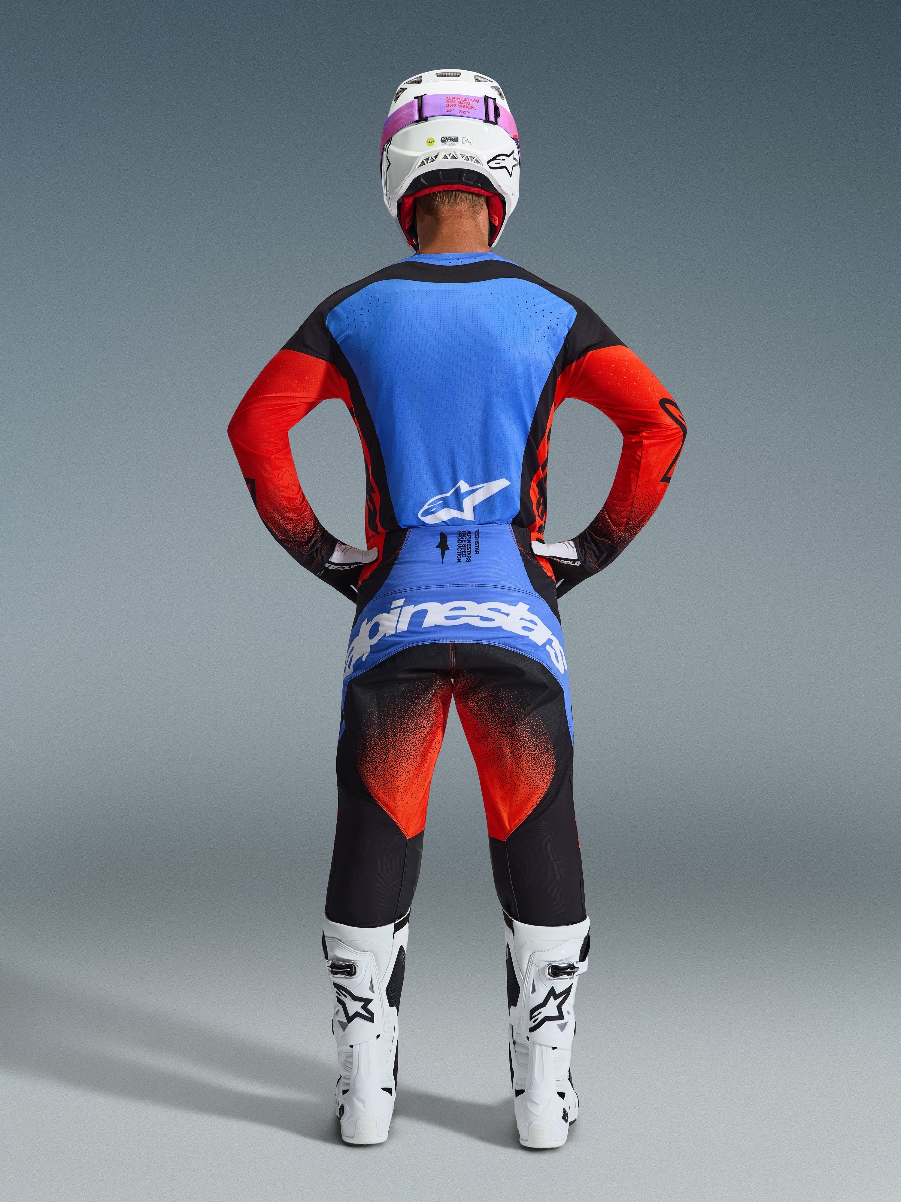 Motocross rider seen from behind wearing the 2026 Techstar Knif Pants in Orange, Black, and UCLA Blue, coordinated with a blue and orange jersey, white helmet with pink goggles, and white racing boots, full Alpinestars off-road competition gear