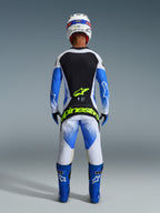 Motocross rider from behind wearing complete racing gear, 2026 Techstar Knif Pants in UCLA Blue Black and Yellow Fluo, matching long-sleeve jersey with black and white panels, blue off-road boots, white helmet, featuring bold fluorescent yellow Alpinestars branding on the seat.