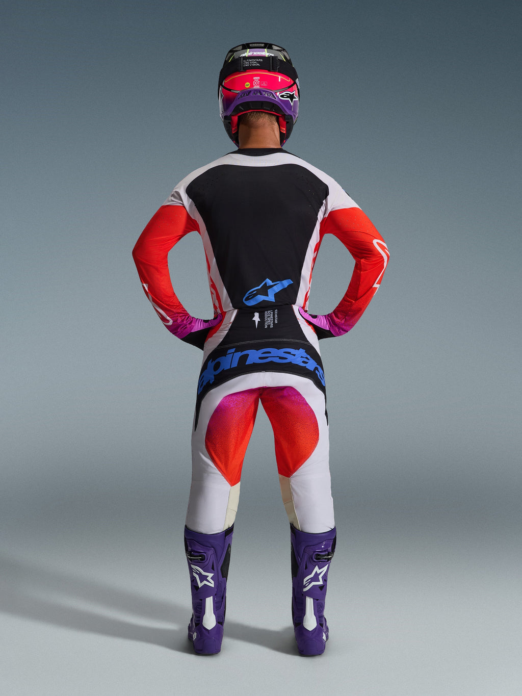 Motocross rider from behind wearing 2026 Techstar Knif Pants and matching jersey, light gray and black design with bright orange and purple accents, blue Alpinestars branding on the seat, purple boots and helmet, professional off-road racing gear set.
