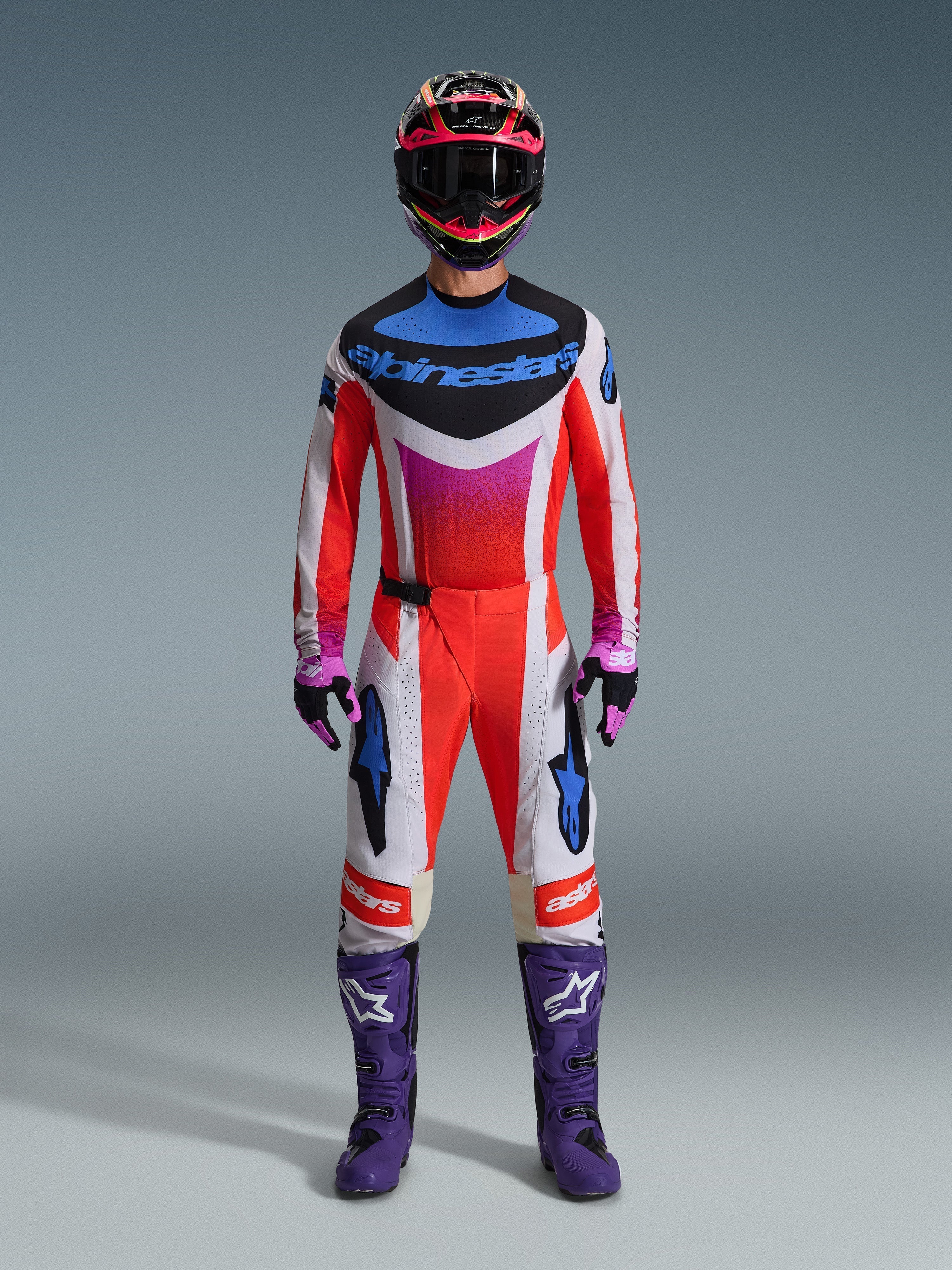 Motocross rider wearing complete racing gear, Alpinestars 2026 Techstar Knif Pants, Fluo orange and white design with blue accents, matching multi-colored jersey, purple boots, black and purple gloves, coordinated professional outfit for motocross racing