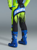 Alpinestars 2026 Techstar Nomur Pants, Motocross Pants, Yellow Fluo and UCLA Blue, rear view of rider wearing blue boots, featuring 'astars' branding on the seat and black splatter-pattern graphics on the legs
