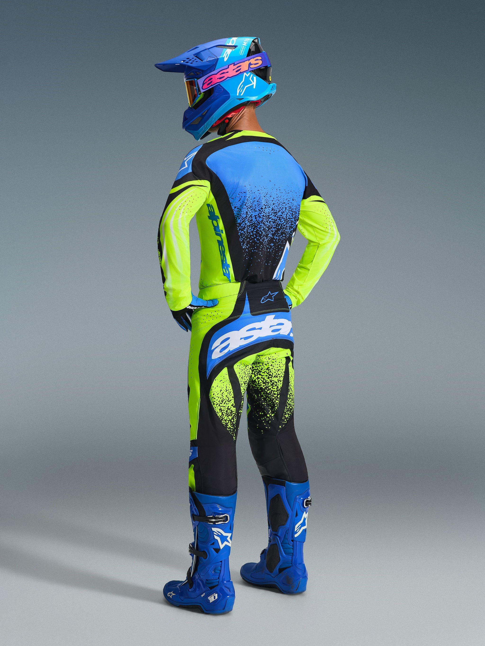 Motocross rider from the back wearing Alpinestars 2026 Techstar Nomur Pants, Yellow Fluo and UCLA Blue, paired with a matching jersey, blue boots, and blue helmet, showing the rear 'astars' logo and black splatter design.