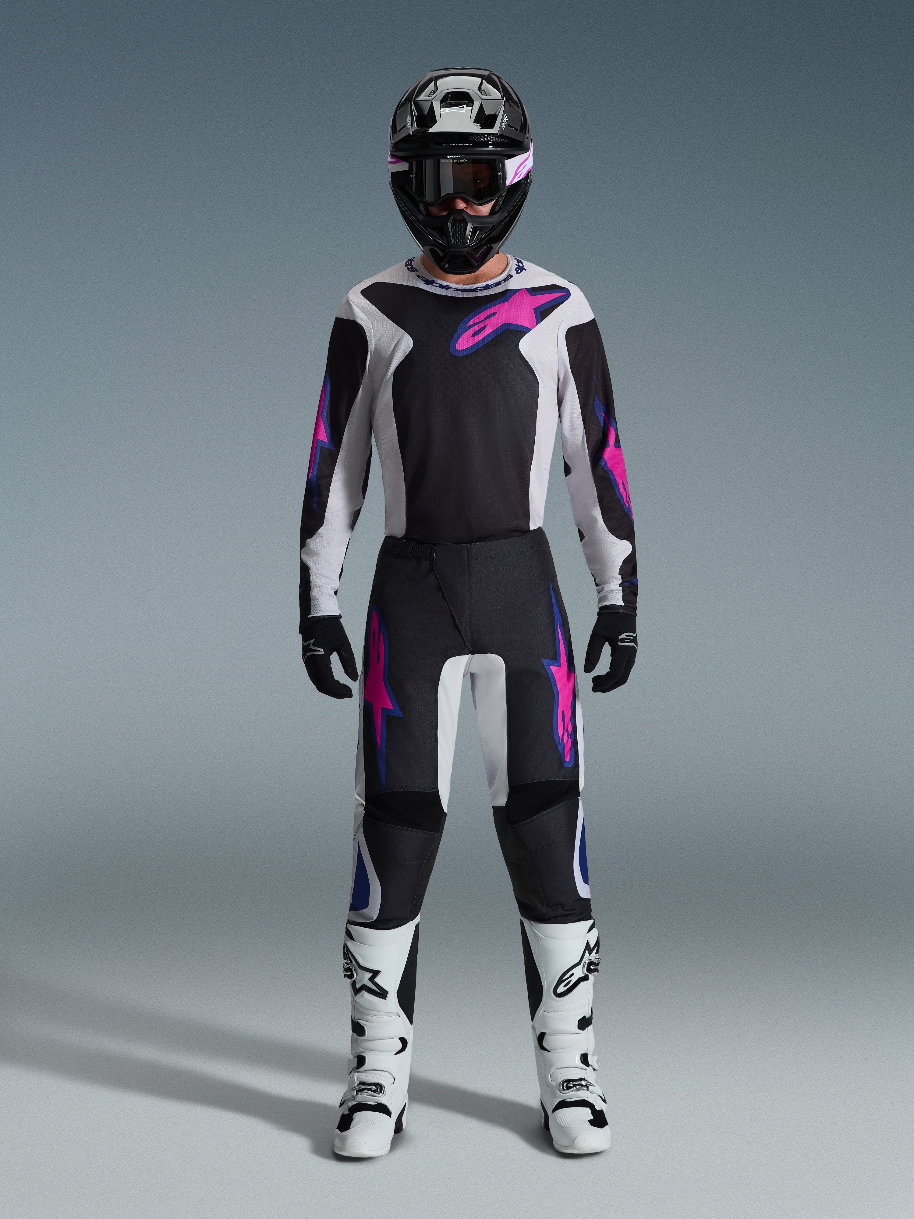 Motocross rider wearing the Alpinestars 2026 Fluid Grid Gear Set, black and light gray jersey with bright pink logos, black and white pants with matching pink and blue graphics, black helmet with pink goggles, white and black boots, full off-road racing gear ensemble