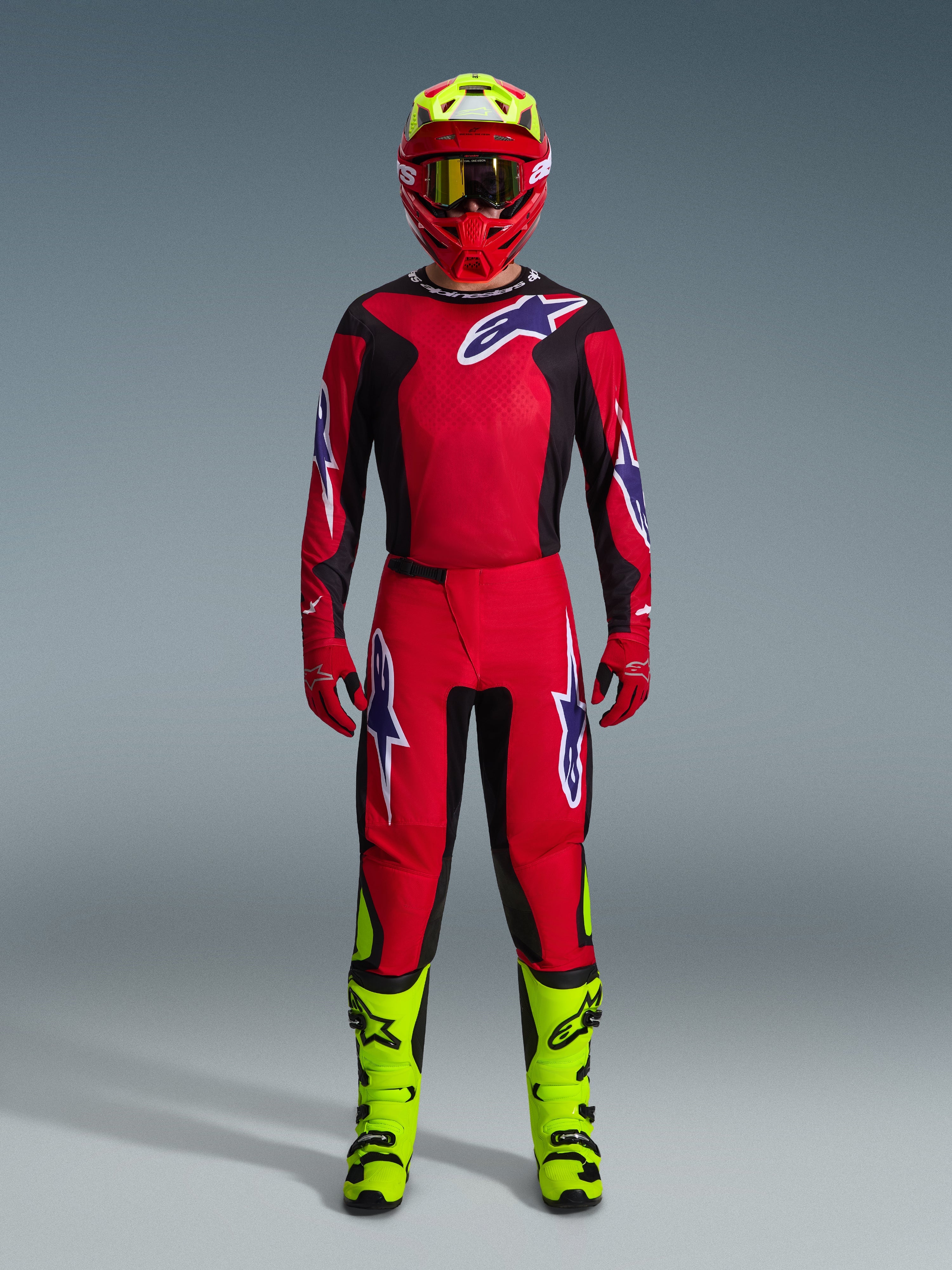 Motocross rider wearing complete racing gear, Alpinestars 2026 Fluid Grid Pants in Red Black and Purple, matching red and black jersey with Alpinestars logo, red helmet with fluorescent yellow accents, fluorescent yellow boots, and red gloves, full gear set for off-road motocross racing