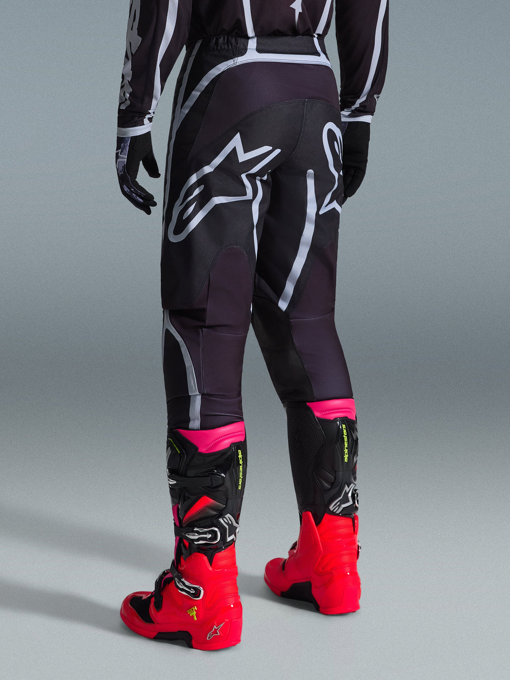 Alpinestars 2026 Fluid Apex Pants, Motocross Pants, Black and Gray, rear view featuring large white Alpinestars logos on the seat, worn with bright red and pink boots, durable 600D polyfabric construction for off-road performance