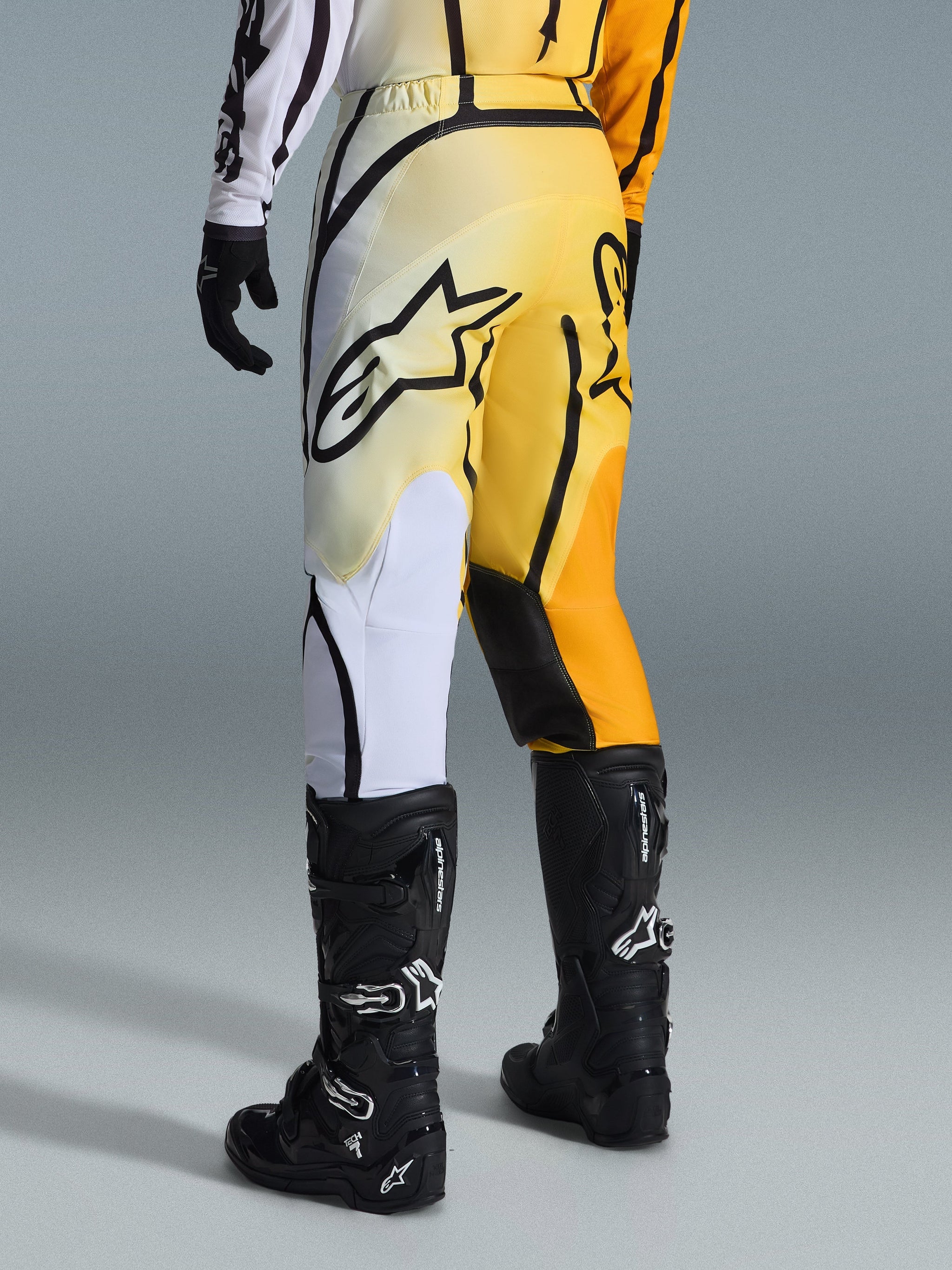 Alpinestars 2026 Fluid Apex Pants, Motocross Pants, Yellow and Black gradient with white and orange accents, viewed from the back, worn with matching jersey and black Tech 7 boots, featuring a bold black Alpinestars logo on the seat, performance gear for off-road riding.