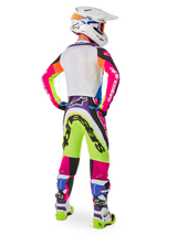 Motocross rider from behind wearing Alpinestars Limited Edition Supertech Hay gear, vibrant multicolor Supertech Hay Pants with neon yellow, hot pink, and purple graphics, matching jersey, white helmet, and coordinated white and blue boots, complete off-road racing outfit