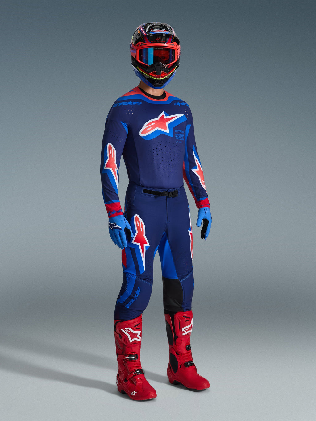 Motocross rider wearing 2026 Supertech Vista Pants in Dark Navy Hot Coral Cobalt, matching long-sleeve racing jersey, red motocross boots, and coordinated helmet with red goggles, full-body studio shot of high-performance off-road gear