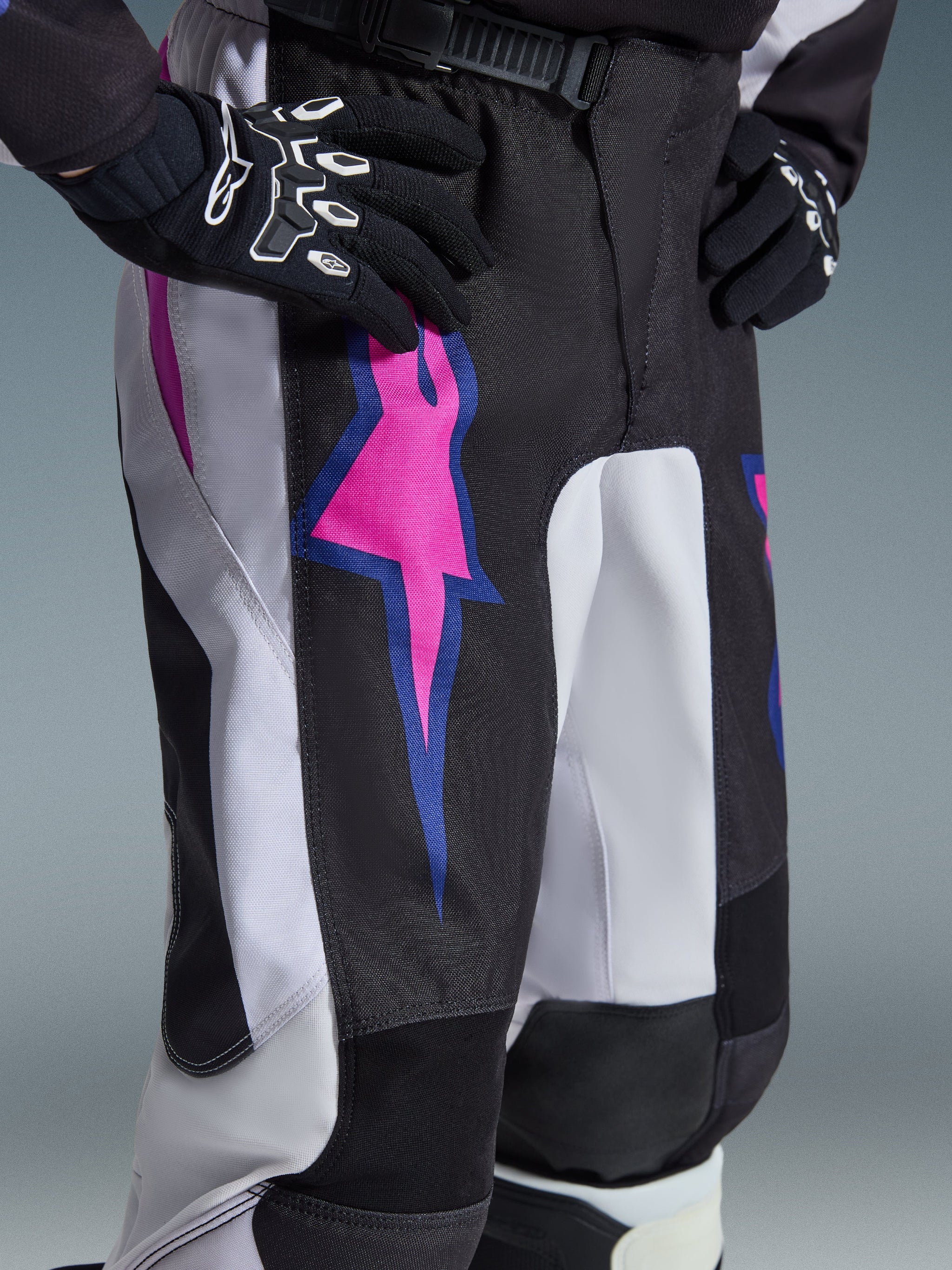 Alpinestars 2026 Youth Fluid Grid Pants, Motocross Pants, Black with Light Gray and Purple, close-up view of the thigh featuring a vibrant pink and blue star graphic, ratchet waist closure, worn with black Alpinestars gloves.