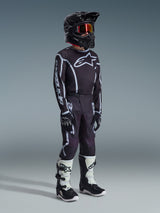 Alpinestars 2026 Youth Fluid Apex Pants, Motocross Pants, Black and Gray, worn with matching black and white jersey, black helmet, and white/black boots, featuring bold Alpinestars branding and graphic lines, coordinated racing outfit for youth motocross