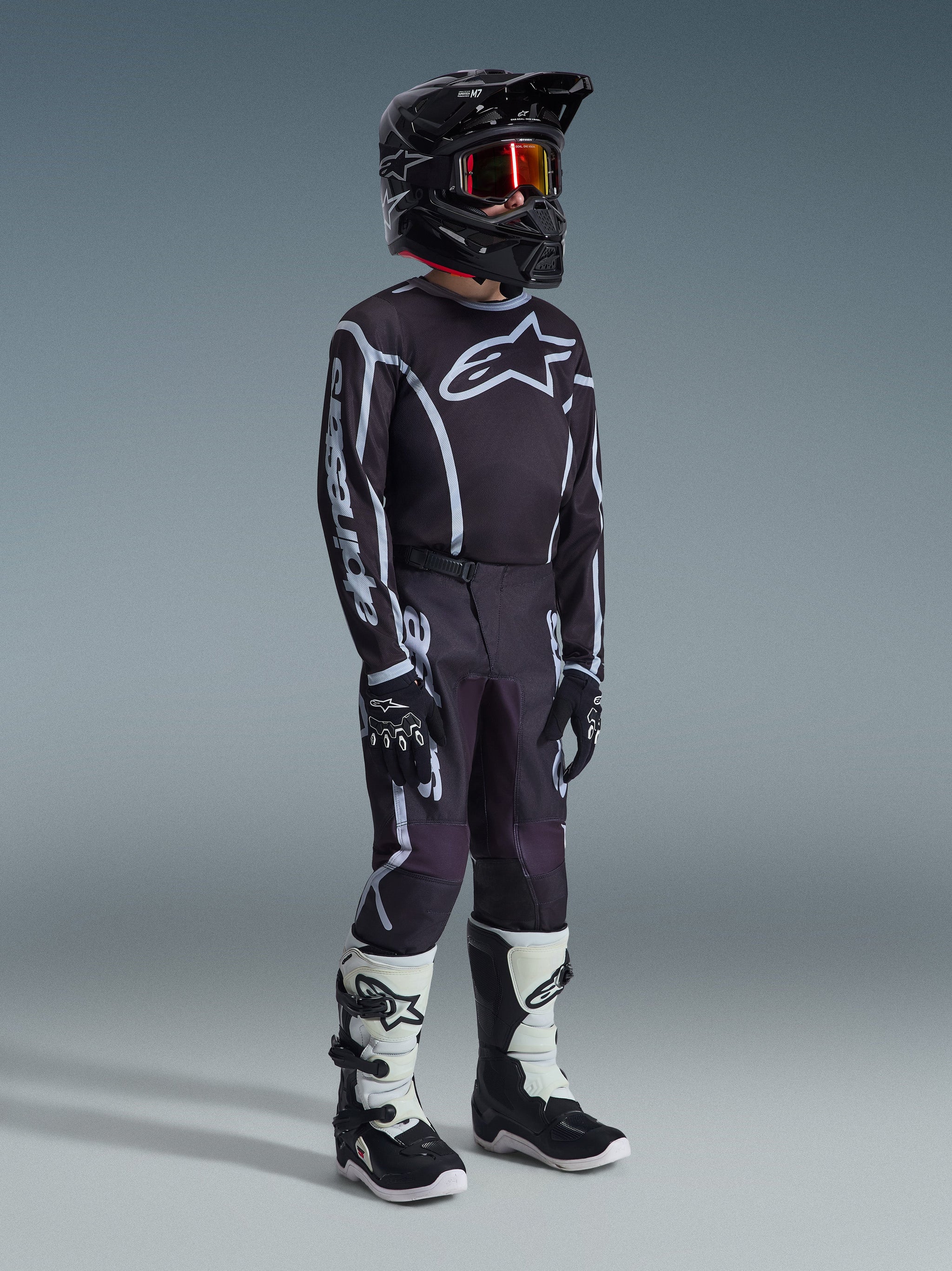 Alpinestars 2026 Youth Fluid Apex Pants, Motocross Pants, Black and Gray, worn with matching black and white jersey, black helmet, and white/black boots, featuring bold Alpinestars branding and graphic lines, coordinated racing outfit for youth motocross