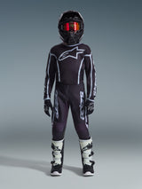 Youth motocross rider wearing complete racing gear, 2026 Youth Fluid Apex Pants in Black and Gray, matching black jersey with white Alpinestars logo, black helmet with red mirrored goggles, white and black boots, black and white gloves, coordinated outfit for off-road racing