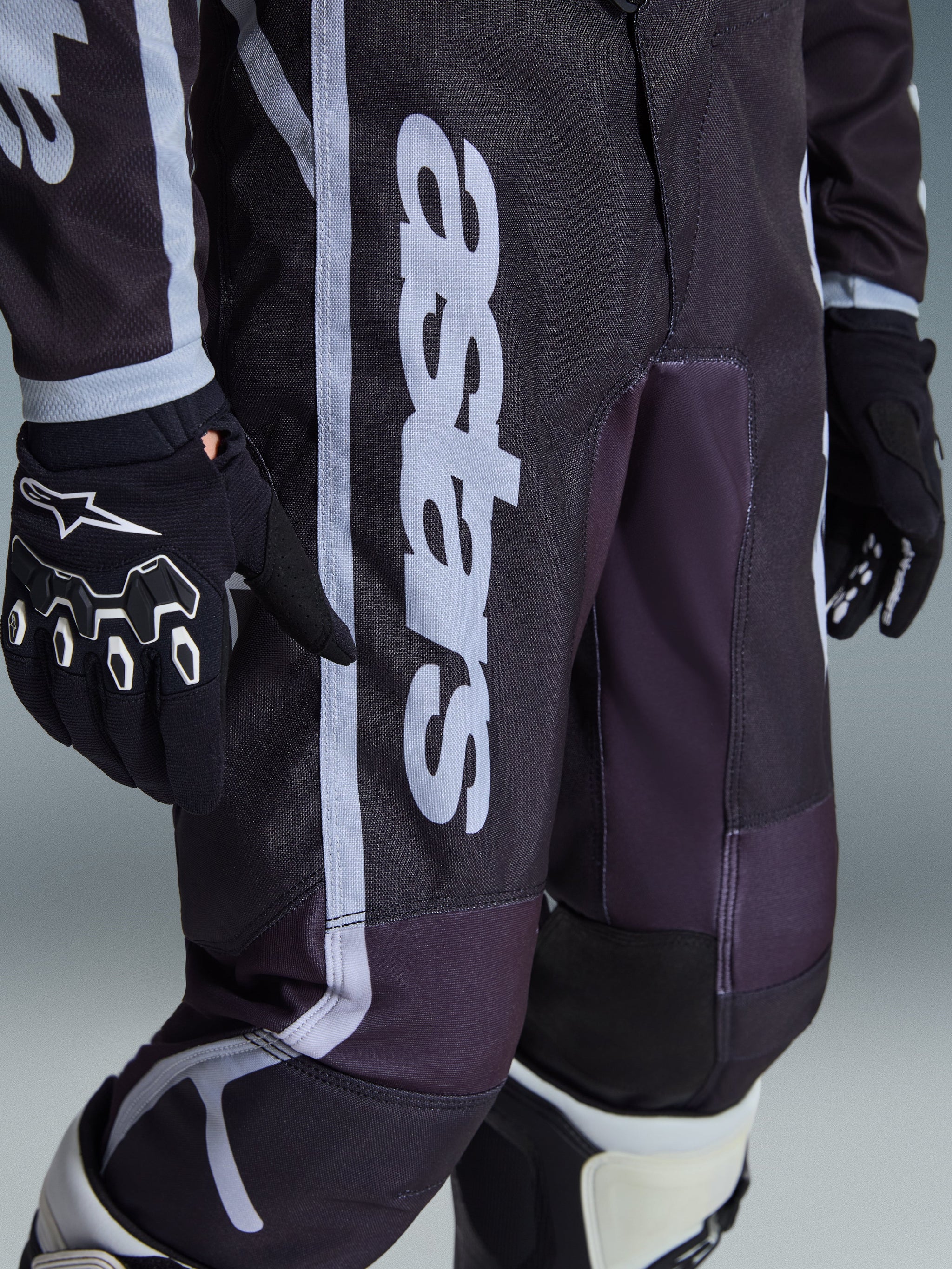 Alpinestars 2026 Youth Fluid Apex Pants, Motocross Pants, Black and Gray, close-up view showing white 'astars' branding on the leg, worn with matching black and white gloves and boots, sporty design for youth off-road racing
