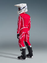 Youth motocross rider wearing complete racing gear from behind, Red and White Alpinestars 2026 Youth Fluid Apex Pants and matching jersey, white helmet, black and white boots, featuring bold white logo graphics and black inner knee panels, coordinated outfit for off-road racing