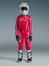 Youth motocross rider wearing complete racing gear, Red jersey with white Alpinestars logo, matching 2026 Youth Fluid Apex Pants in Red and White, white helmet and goggles, white and black boots, black and white gloves, coordinated outfit for off-road racing
