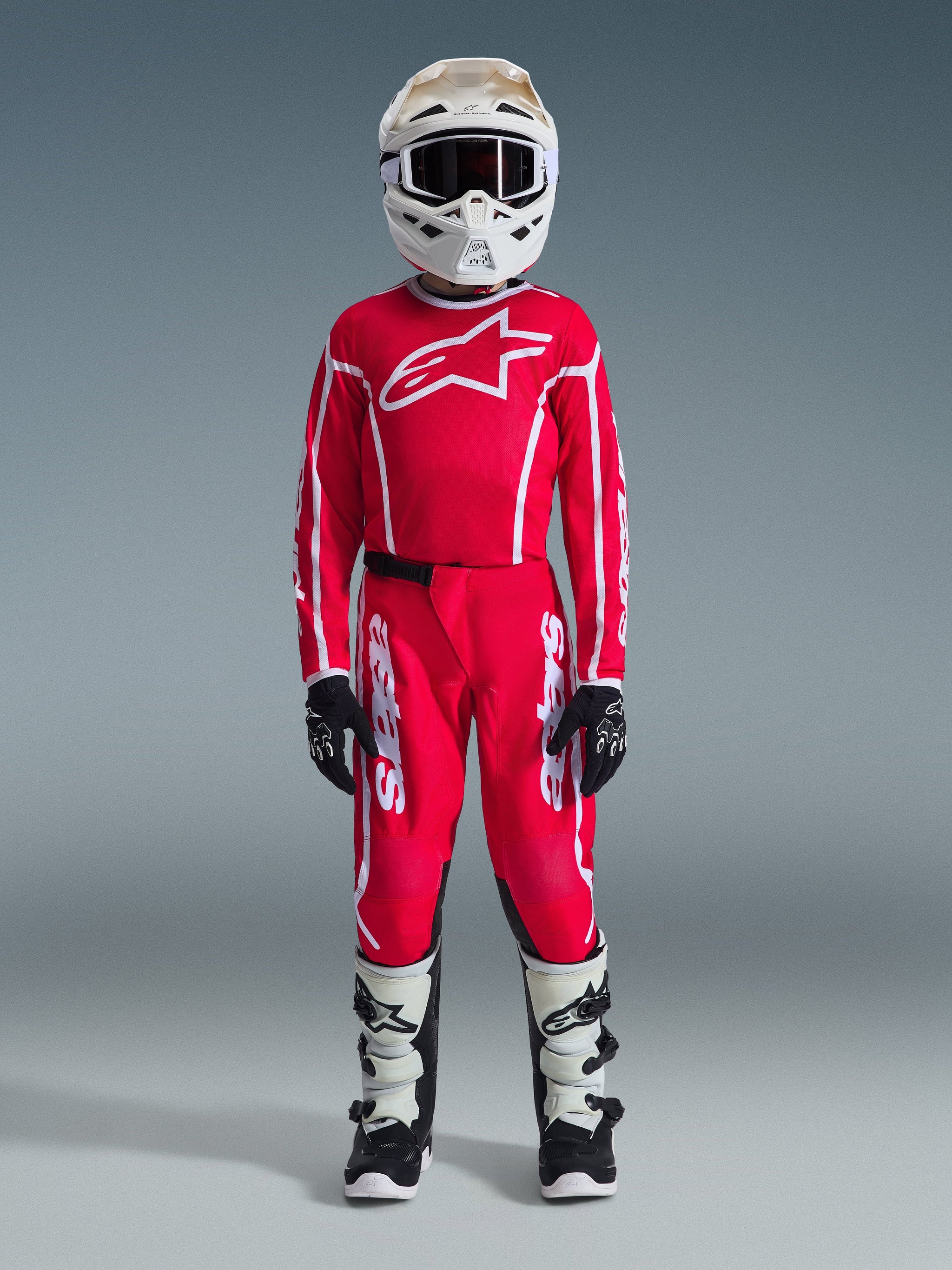 Youth motocross rider wearing complete racing gear, Red jersey with white Alpinestars logo, matching 2026 Youth Fluid Apex Pants in Red and White, white helmet and goggles, white and black boots, black and white gloves, coordinated outfit for off-road racing