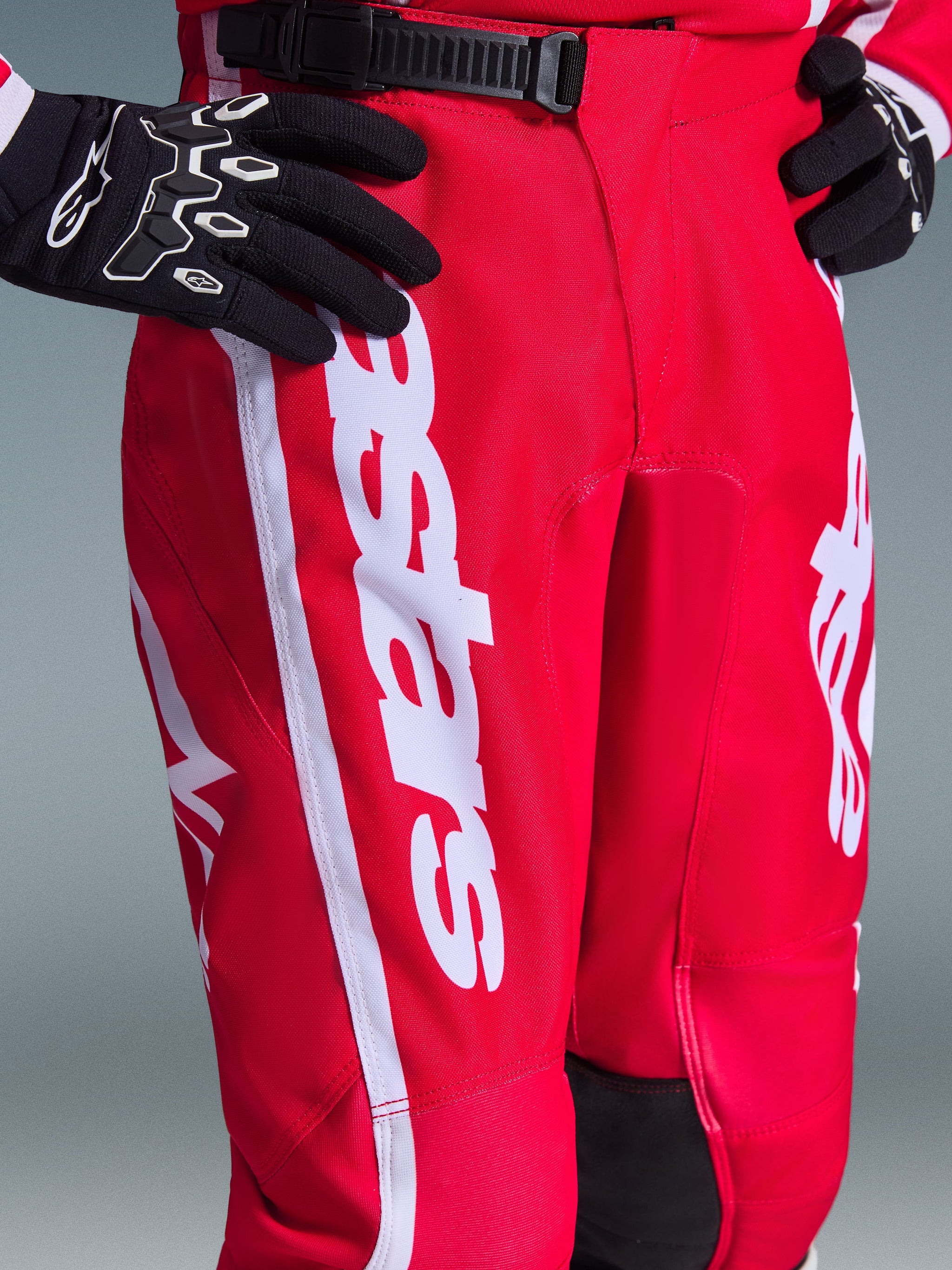 Alpinestars 2026 Youth Fluid Apex Pants, Motocross Pants, Red and White, featuring black ratchet waist closure and bold white Alpinestars branding on the thigh, worn with black and white gloves, durable polyfabric for off-road racing