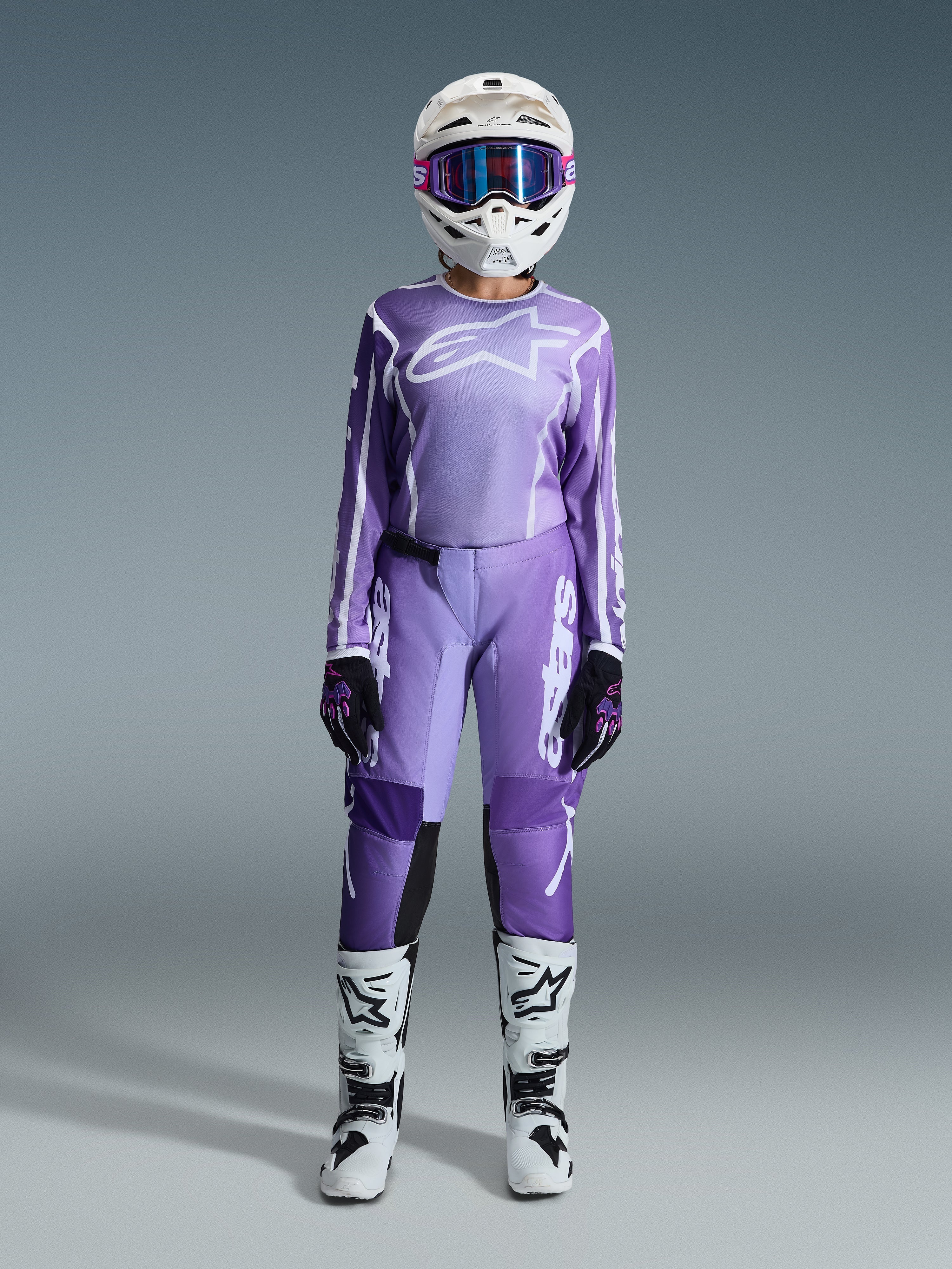 Motocross rider wearing the Alpinestars 2026 Woman Stella Fluid Apex Gear Set, violet and white jersey with matching pants, white helmet with pink goggles, and white and black off-road boots, complete racing kit for women's motocross.