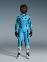 Female motocross rider wearing the 2026 Alpinestars Stella Fluid Apex gear set, bright cyan blue jersey and pants with black accents, black helmet with iridescent goggles, and white boots, full professional motocross outfit