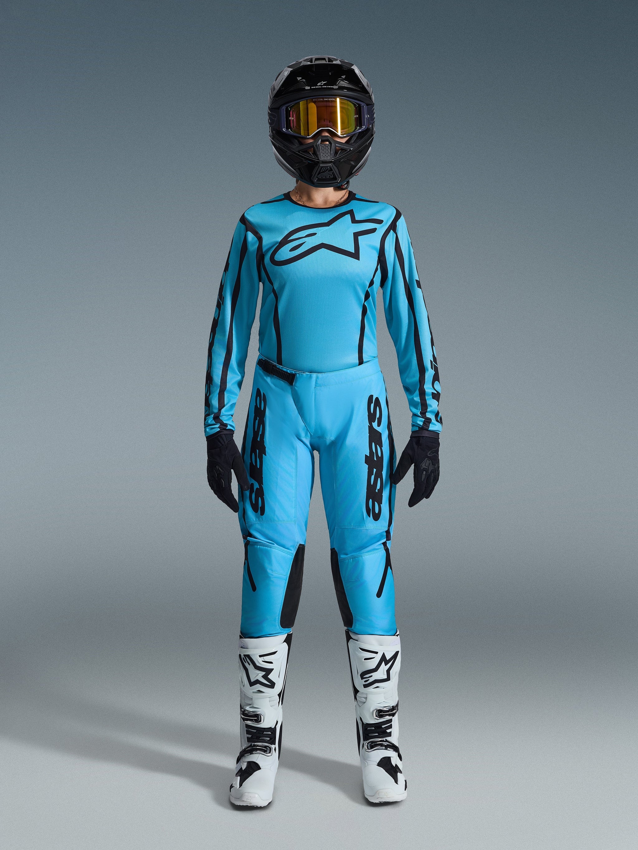 Female motocross rider wearing the 2026 Alpinestars Stella Fluid Apex gear set, bright cyan blue jersey and pants with black accents, black helmet with iridescent goggles, and white boots, full professional motocross outfit