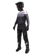 Alpinestars Pro-Dura Jersey in Black Magnet White, worn by a motocross rider in full gear including black pants, black helmet with goggles, and black and white boots. The jersey features a technical color-blocked design with white, grey, and black panels, engineered for enduro and off-road performance.