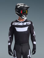 Motocross rider wearing the Alpinestars 2026 Racer Air Riway Gear Set, black jersey with white 'astars' logo and ventilated chest panel, black pants with white accents, matching black and white helmet with iridescent goggles, black gloves, high-performance motocross gear in black and white.
