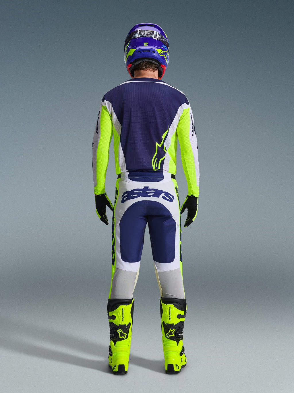 Motocross rider from behind wearing the Alpinestars 2026 Racer Air Riway Gear Set, Blue jersey with fluo yellow and light gray accents, white and blue pants with fluo yellow side panels, fluo yellow boots with black details, matching blue and yellow helmet, professional gear for warm-weather off-road riding