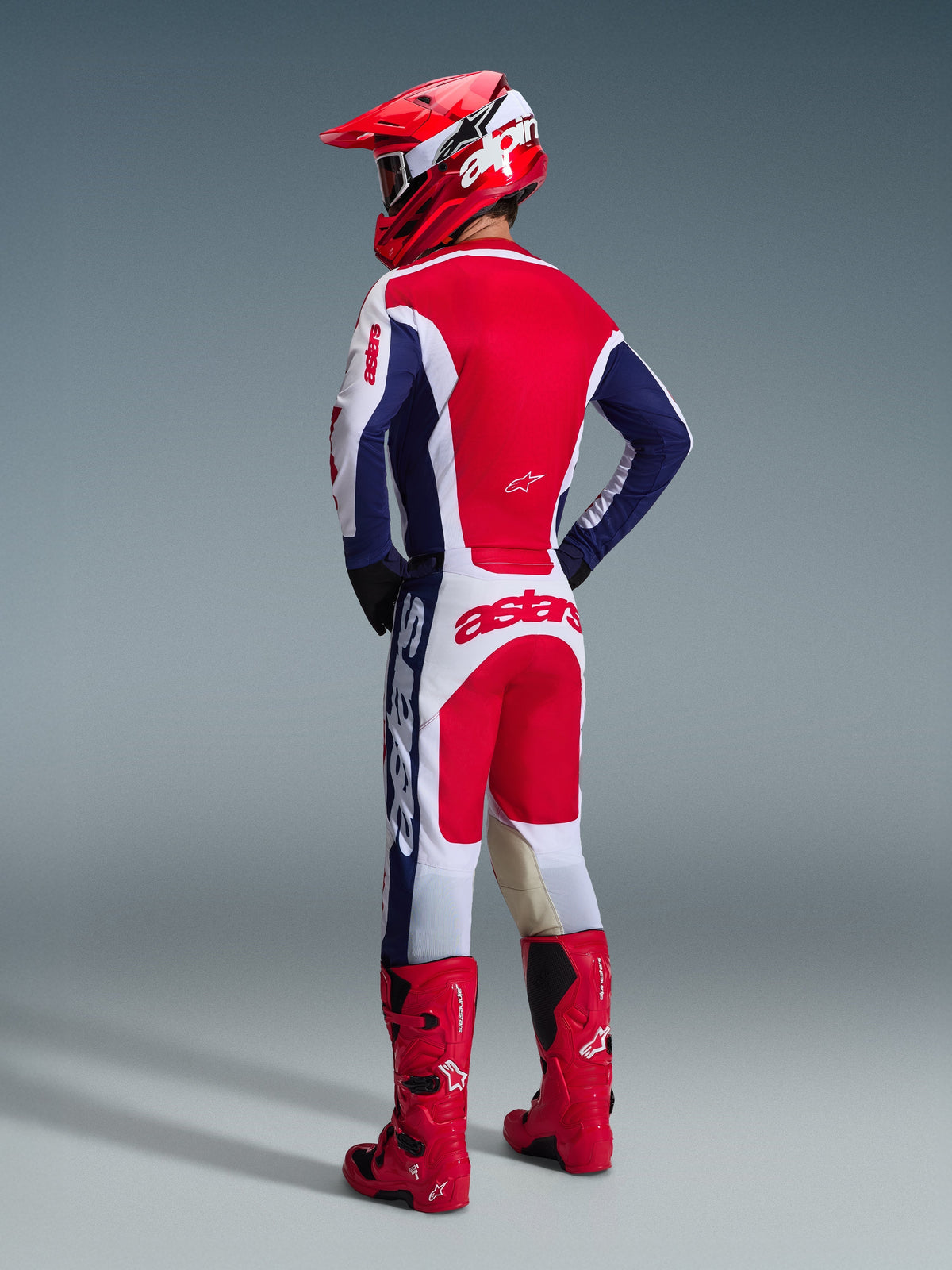 Motocross rider from behind wearing Alpinestars 2026 Racer Riway Jersey in White Blue Red, matching motocross pants with 'astars' branding on the seat, red and white helmet, and red motocross boots, complete off-road racing gear