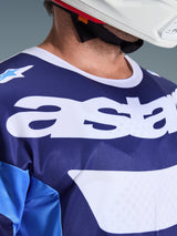 Close-up of Alpinestars 2026 Racer Riway Jersey in Ucla Blue and White, featuring large white 'astars' logo on the chest, white collar, and breathable mesh fabric texture, worn with a white motocross helmet