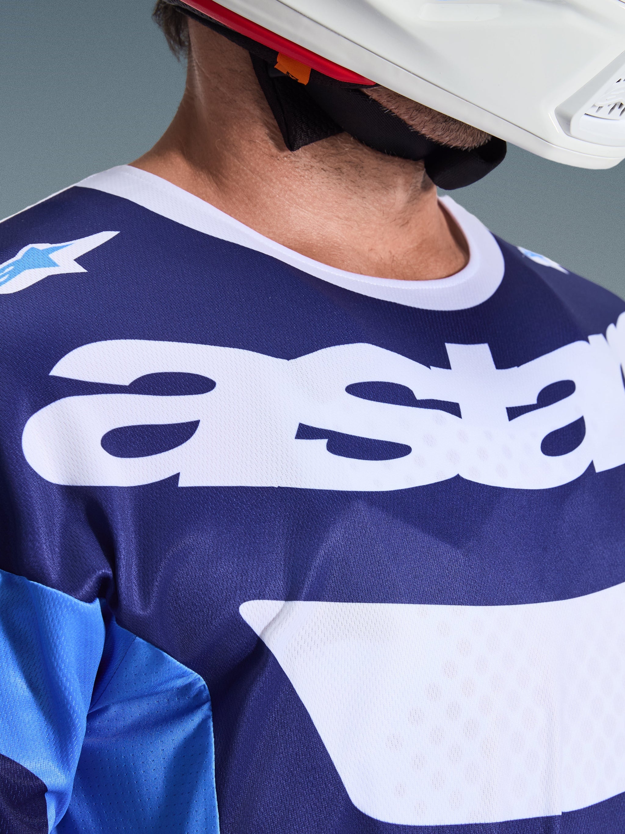 Close-up of Alpinestars 2026 Racer Riway Jersey in Ucla Blue and White, featuring large white 'astars' logo on the chest, white collar, and breathable mesh fabric texture, worn with a white motocross helmet