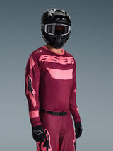 Motocross rider wearing the Alpinestars 2026 Racer Riway Jersey in Dark Red Coral with matching pants, featuring coral pink 'astars' branding on the chest, paired with a black helmet and black gloves, coordinated off-road racing gear set.