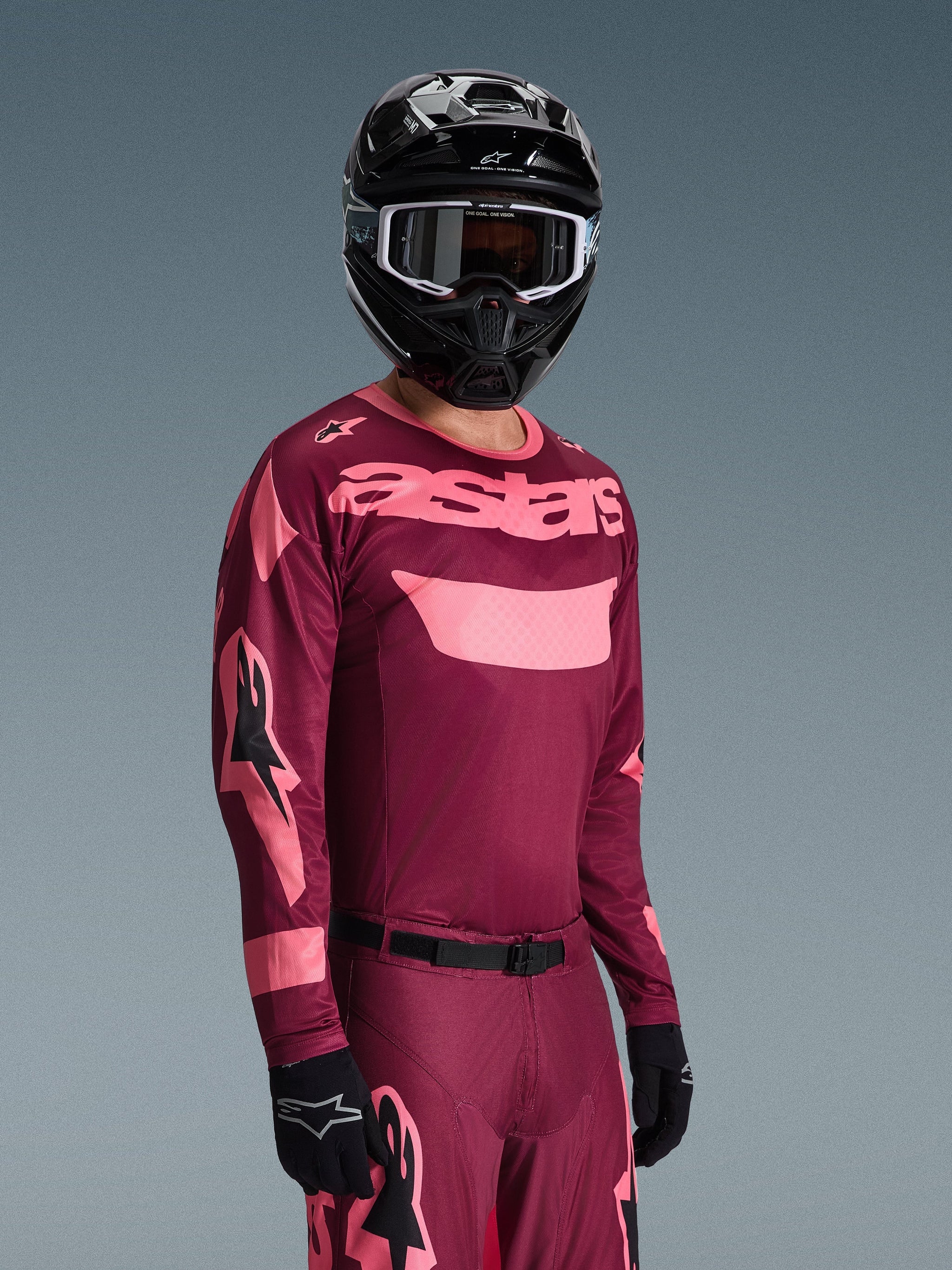 Motocross rider wearing the Alpinestars 2026 Racer Riway Jersey in Dark Red Coral with matching pants, featuring coral pink 'astars' branding on the chest, paired with a black helmet and black gloves, coordinated off-road racing gear set.