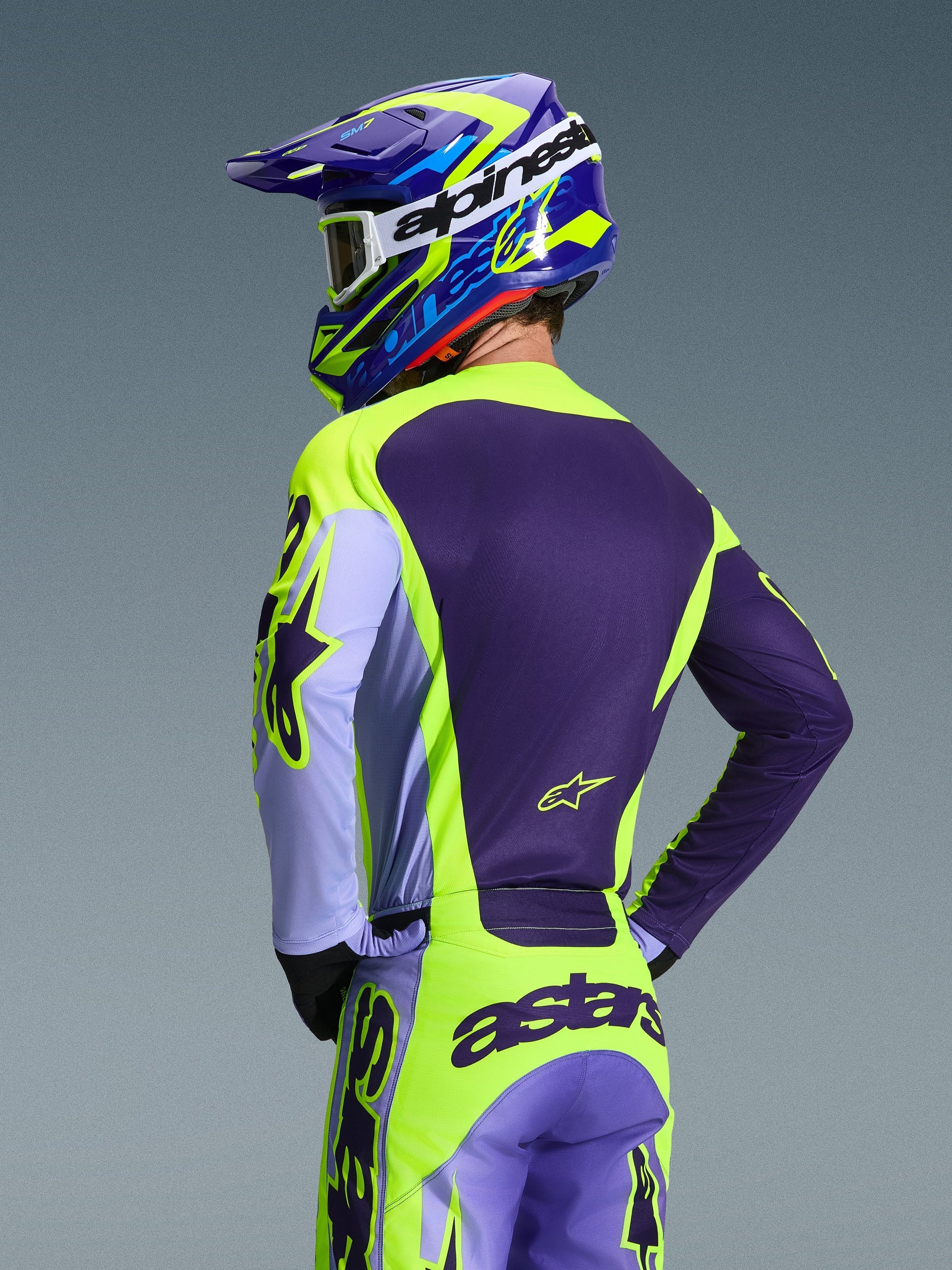 Motocross rider from behind wearing the Alpinestars 2026 Racer Portl Jersey and matching pants, Purple and Yellow Fluo color scheme with lavender accents, coordinated blue and yellow helmet, race-fit gear designed for off-road performance.