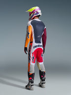 Motocross rider from behind wearing the Alpinestars 2026 Racer Portl Gear Set, featuring a color-blocked jersey with orange and maroon sleeves, white pants with red panels and bold 'astars' branding, multi-colored helmet, and gray boots, professional off-road racing outfit.