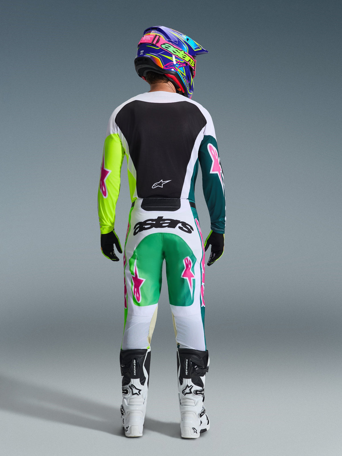 Motocross rider viewed from the back wearing the 2026 Racer Portl Jersey in Green White Black, featuring a black back panel and asymmetric green sleeves with pink star logos, styled with matching motocross pants and white boots.
