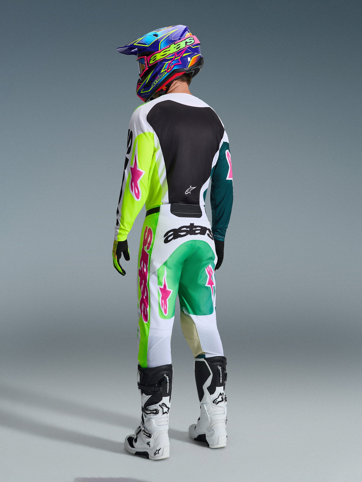 Motocross rider wearing the Alpinestars 2026 Racer Portl Jersey in Green White Black, seen from behind, matching racing pants with pink star logos and 'astars' branding, white and black boots, and a colorful helmet, complete coordinated outfit for off-road racing