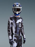 Motocross rider wearing the Alpinestars 2026 Racer Lahnd Jersey in Iron Camo, featuring a black and grey camouflage pattern with a large white Alpinestars logo on the chest, styled with a matching helmet and gloves for off-road racing.