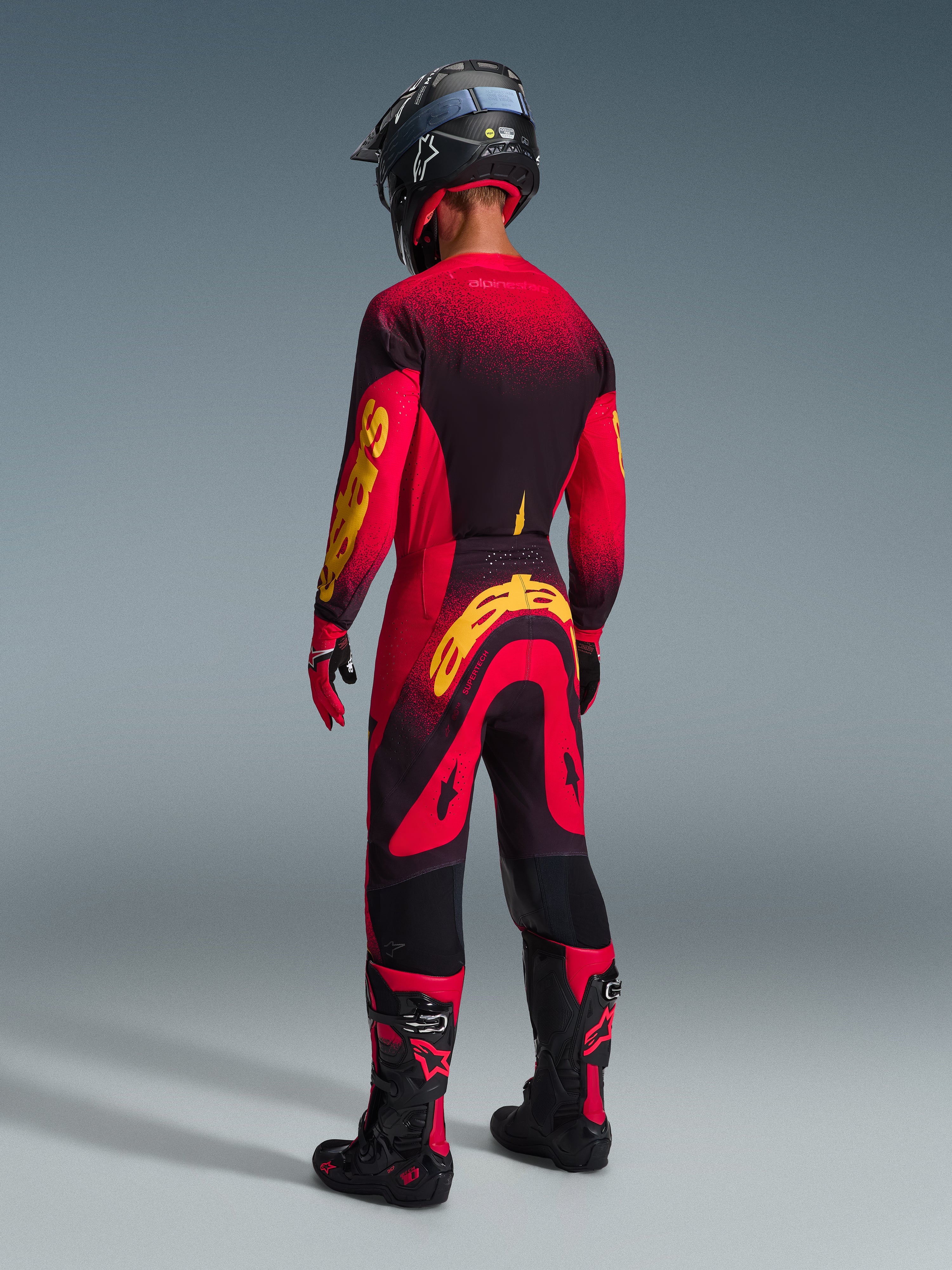 Alpinestars 2026 Supertech Scenz Gear Set, motocross rider viewed from behind, red and black jersey with yellow branding, matching red and black pants with yellow logo, red gloves, black and red motocross boots, and black helmet, coordinated pro-level racing gear.