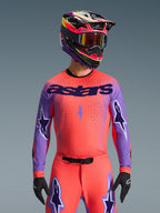 Motocross rider wearing the Alpinestars 2026 Supertech Scenz Gear Set, Vibrant pinkish-orange jersey with purple sleeves and 'astars' branding, matching pants with purple star accents, multi-colored racing helmet with goggles, black gloves, professional off-road racing gear