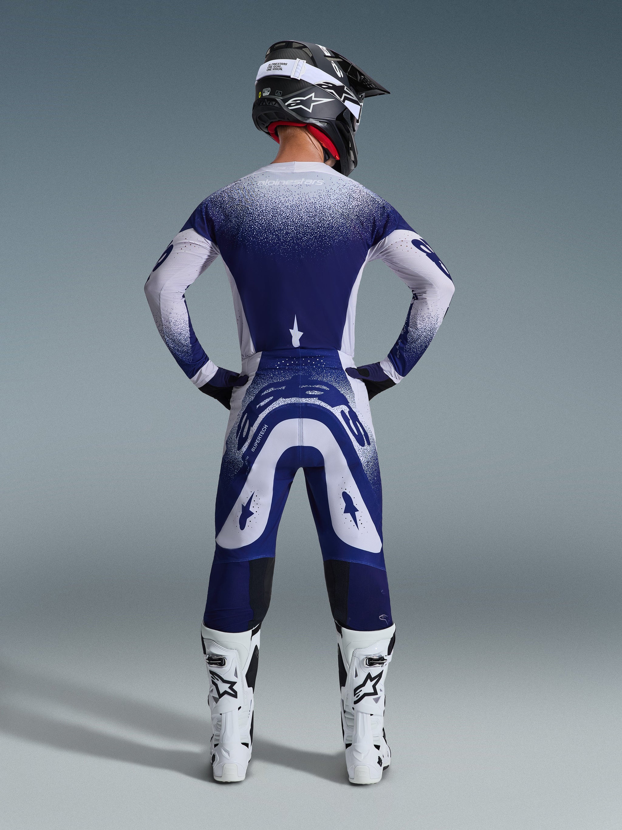 Motocross rider from behind wearing the Alpinestars 2026 Supertech Scenz Jersey, Light Gray and Dark Blue with a stippled gradient design, matching motocross pants, white racing boots, and black helmet, complete off-road racing outfit.