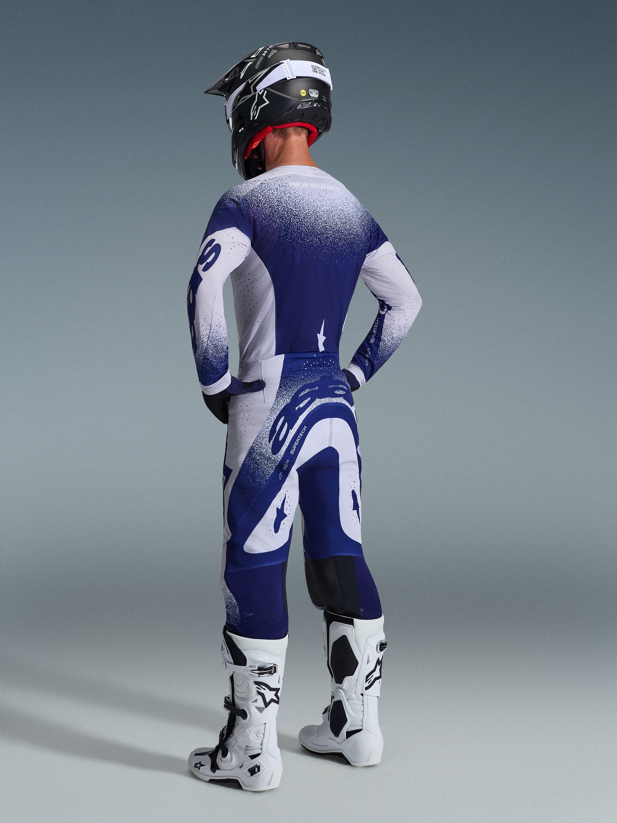 Rear view of a motocross rider wearing the Alpinestars 2026 Supertech Scenz Jersey in Light Gray and Dark Blue, featuring a speckled gradient design, coordinated with matching pants and white motocross boots, professional athletic racing gear.