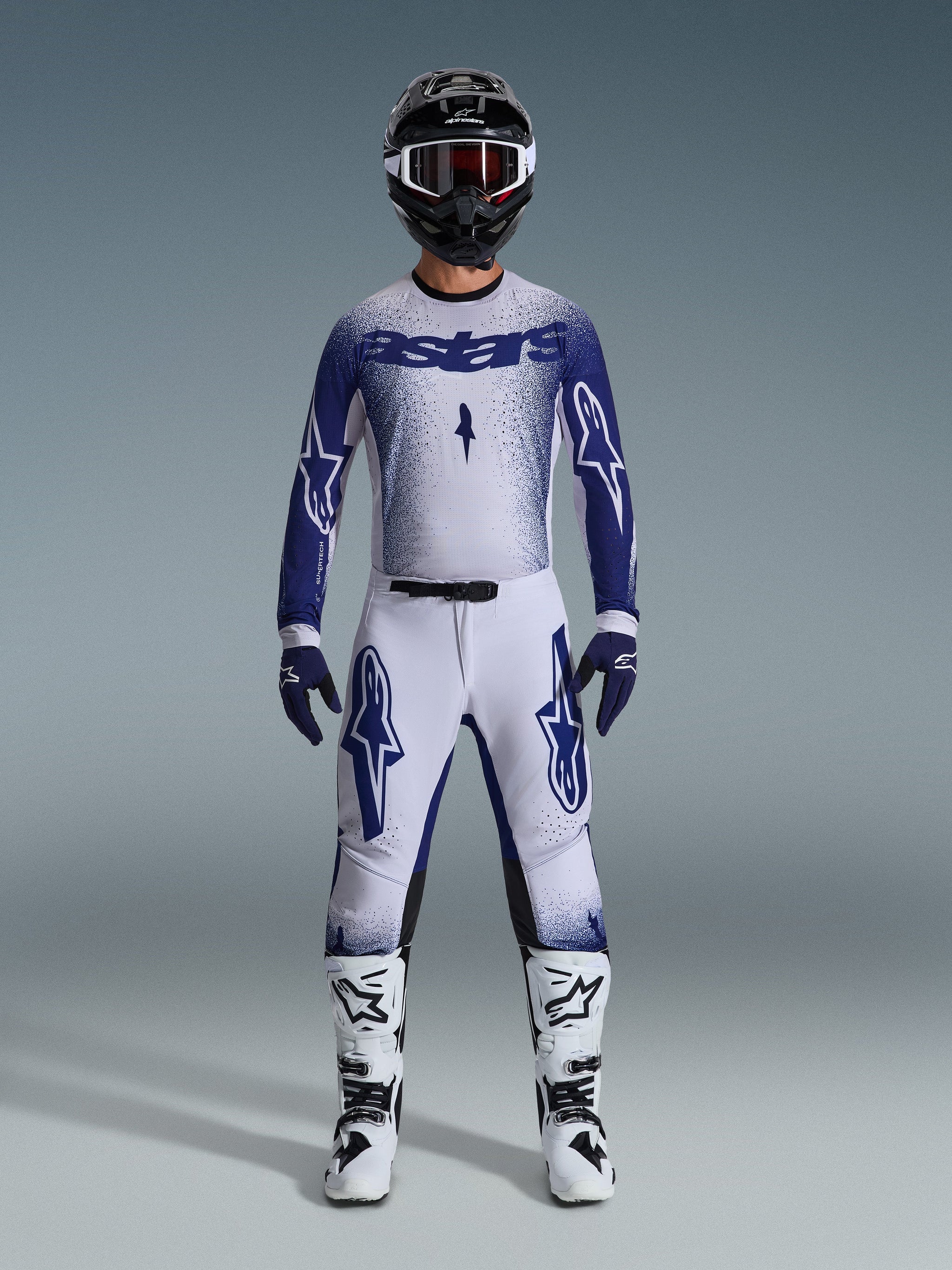 Motocross rider wearing complete racing gear, 2026 Supertech Scenz Jersey in light gray and dark blue, matching light gray pants with blue logos, black and white helmet with red goggles, white and black boots, dark blue gloves, coordinated outfit for off-road racing