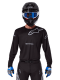Alpinestars 2025 Racer Graphite Jersey, Motocross Racing Gear, Black with Gray logos, worn with matching black helmet, blue goggles, and blue and gray gloves, form-fitting performance design with chest and arm branding