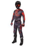 Motocross rider wearing complete racing gear, Alpinestars 2025 Racer Hollow Jersey and Pants in Gray Camo Orange Fluo, black helmet with orange goggles, black and red gloves, and black boots with white logos, coordinated performance outfit for off-road racing