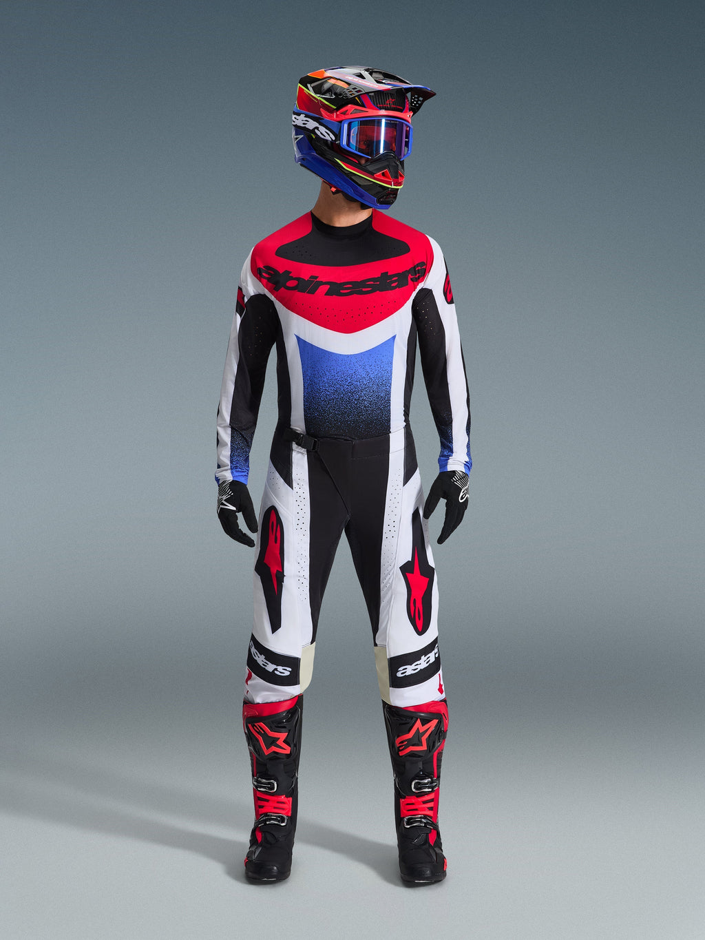 Motocross rider wearing the Alpinestars 2026 Techstar Knif Gear Set, black white and red jersey with blue gradient and branding, matching black and white pants with red accents, multi-colored helmet with blue goggles, and black and red boots, coordinated off-road racing outfit.