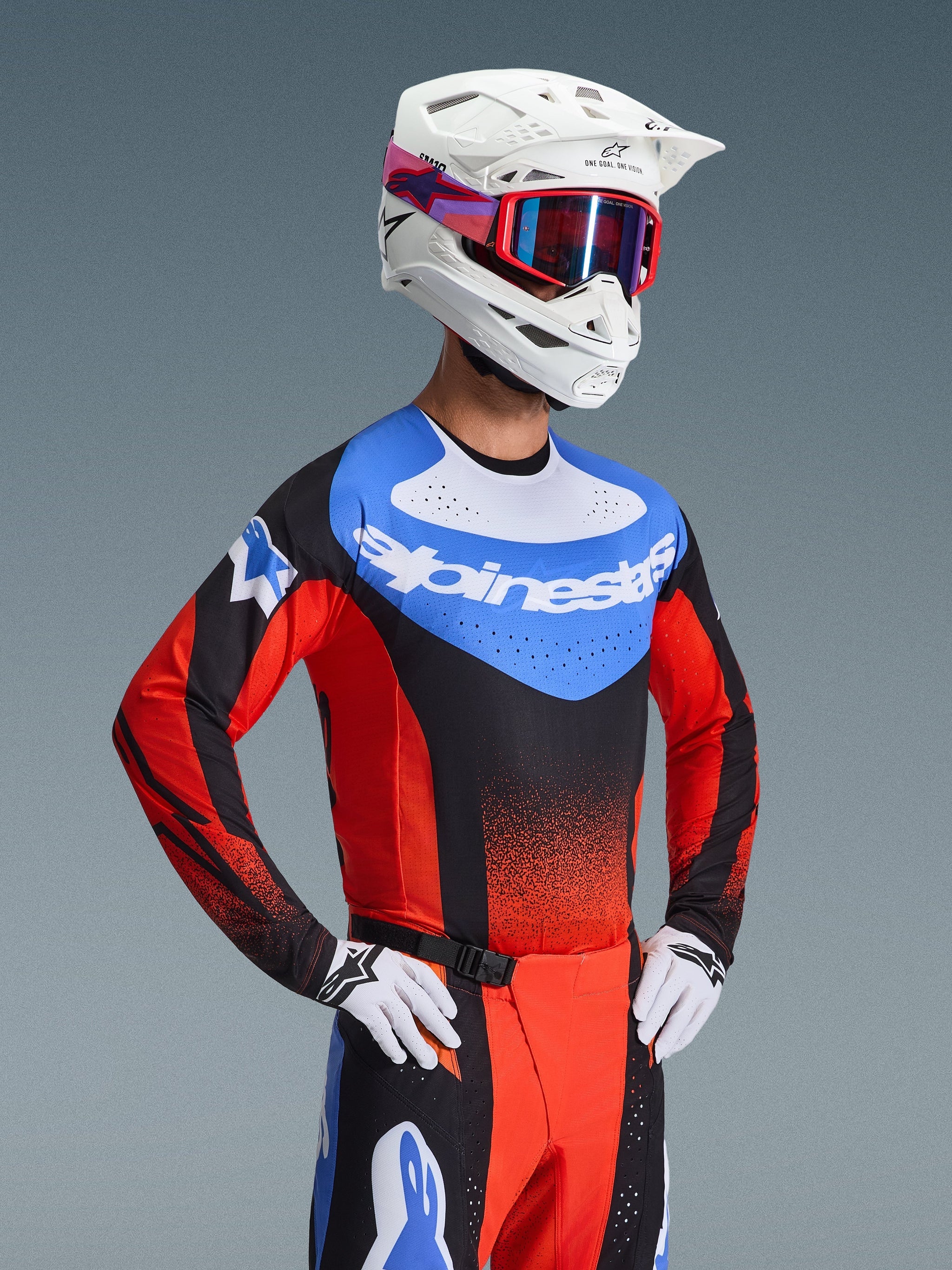 Motocross rider wearing the 2026 Techstar Knif Jersey in Orange Black and UCLA Blue, featuring a blue chest panel with white Alpinestars logo, black and orange sleeves with laser-cut perforations, paired with a white helmet, red goggles, and matching racing pants.